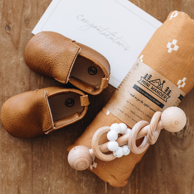 Luxury gift box for a new mom. Includes Sea Mineral Body Polish, size 2 leather baby shoes, a wooden baby rattle, a gold hair claw, and a swaddle, available in orange with boho squares or white with sage green leaves. This all comes packaged in a wooden keepsake gift box.