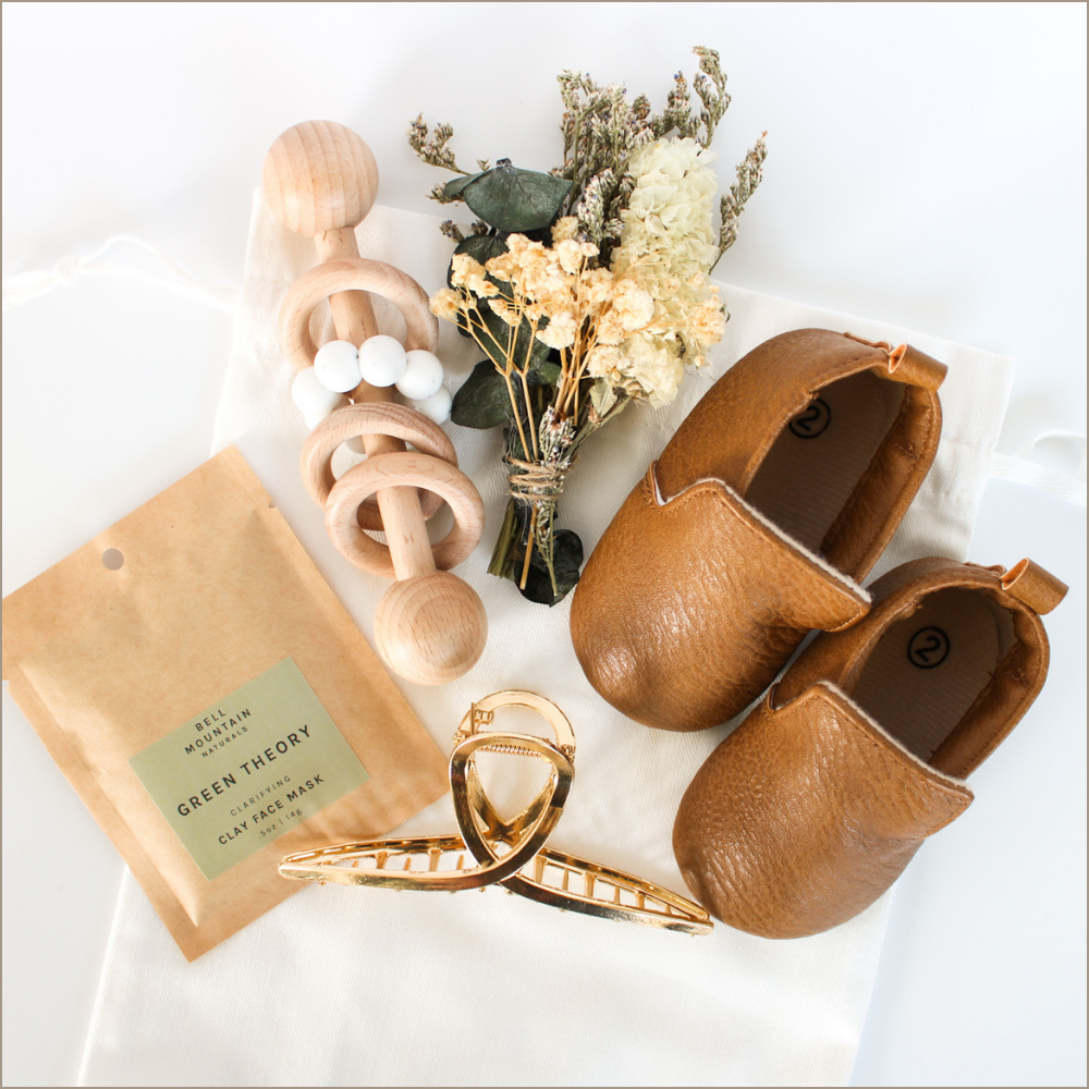 Leather size 2 baby shoes, dried mini flower bouquet, wooden baby rattle, Green Theory clay face mask, gold hair claw all packaged in an organic cotton drawstring gift bag.